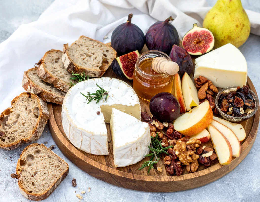 Un plateau élégant de fromages de chèvre artisanaux Pampilles & Compagnie, entouré de tranches de pain rustique, de quartiers de pommes et de poires, de figues fraîches, de noix, d’un petit pot de miel au centre et d’un ramequin de fruits secs.